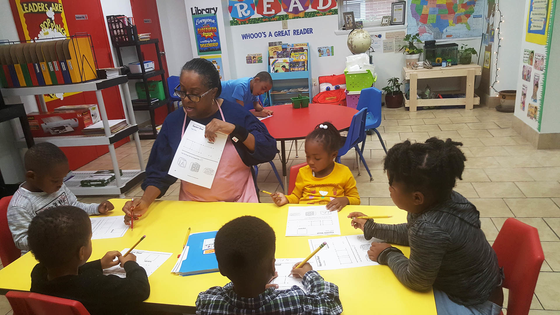 A teacher assisting a small group of children with educational worksheets at a table in a classroom.