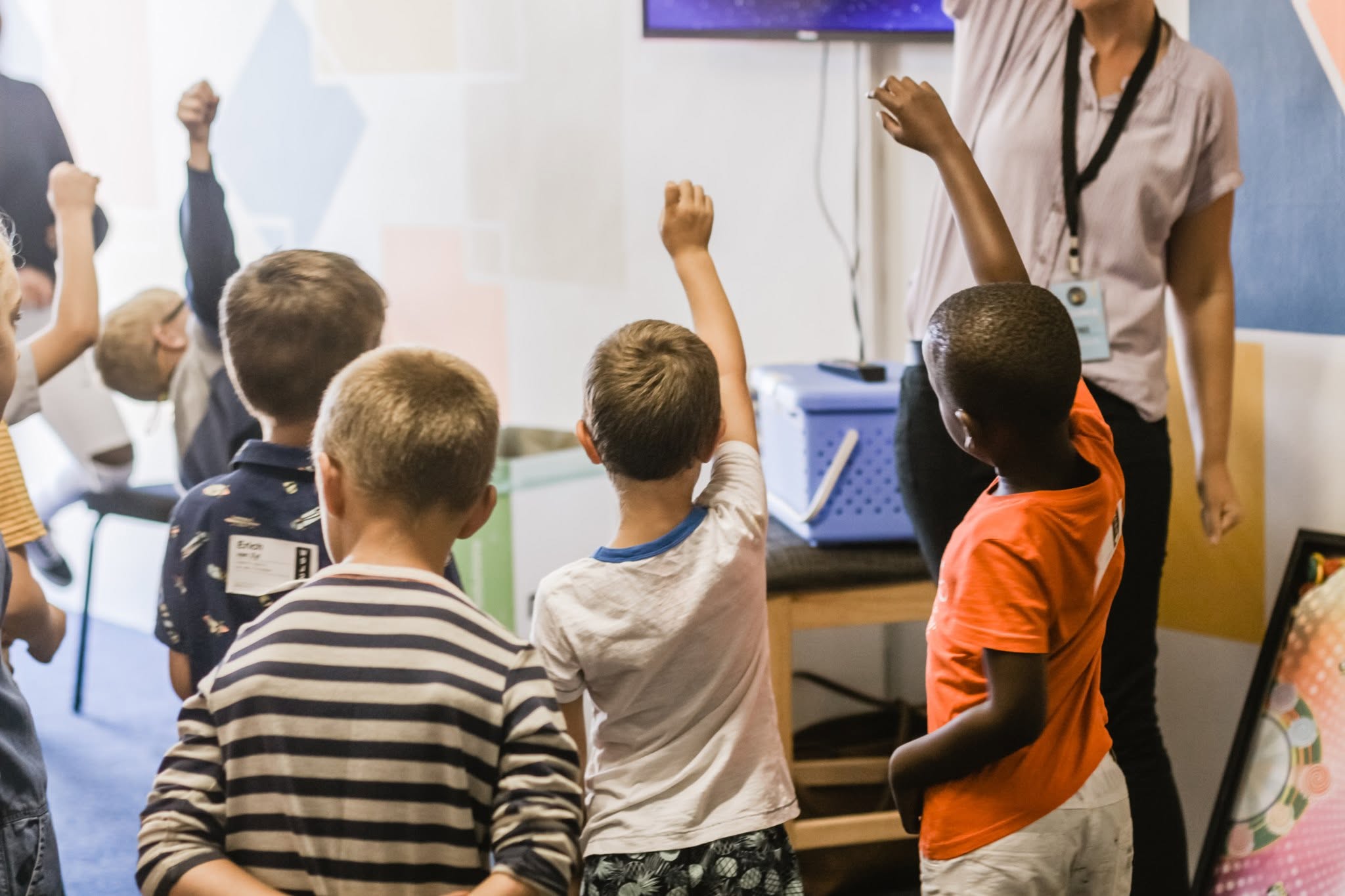 A group of diverse elementary students with their hands raised in a classroom setting.