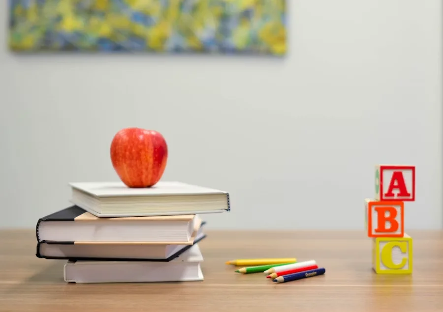 A red apple resting on a stack of books, accompanied by colored pencils and ABC blocks on a wooden table.