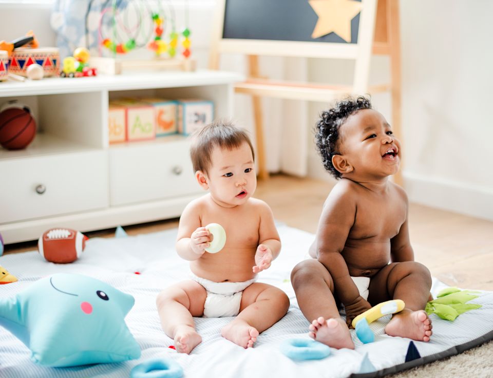 Two diverse babies sitting on a play mat with toys in a childcare setting.