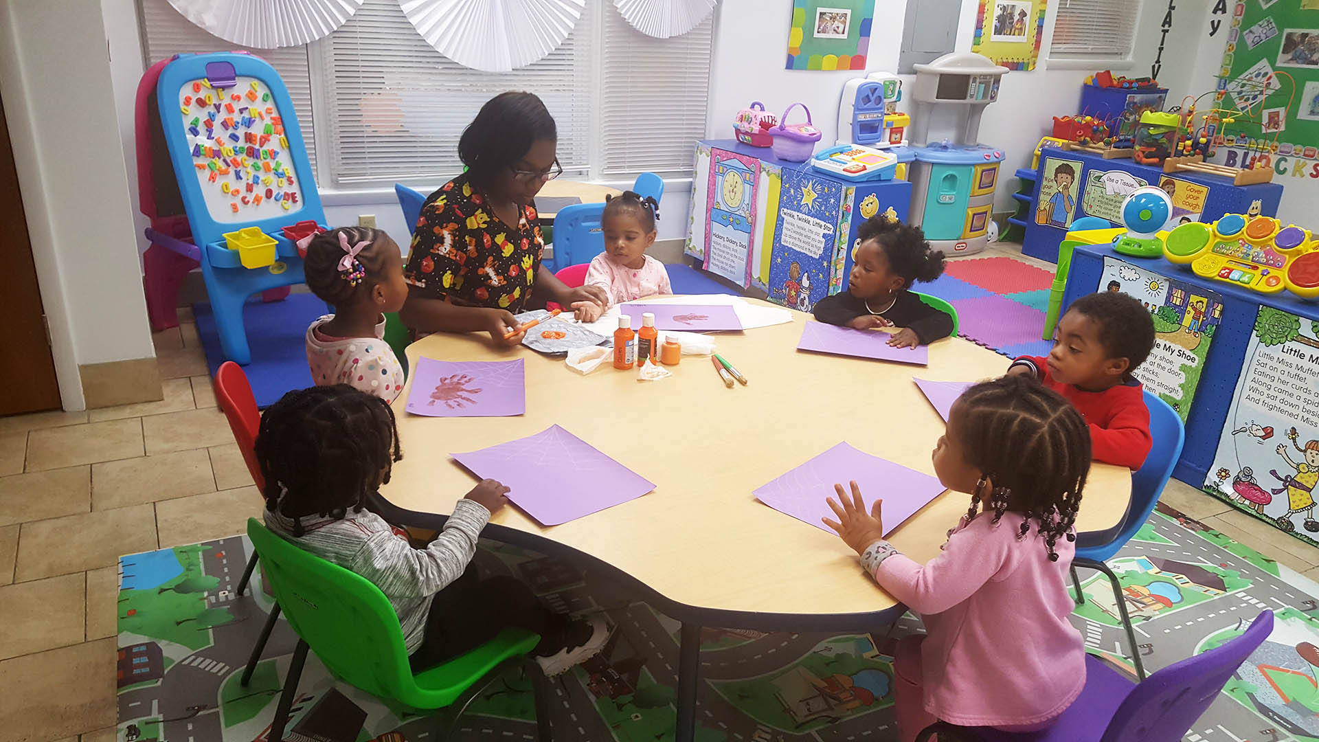 A teacher helping a group of toddlers with an arts and crafts project at a circular table in a daycare setting.